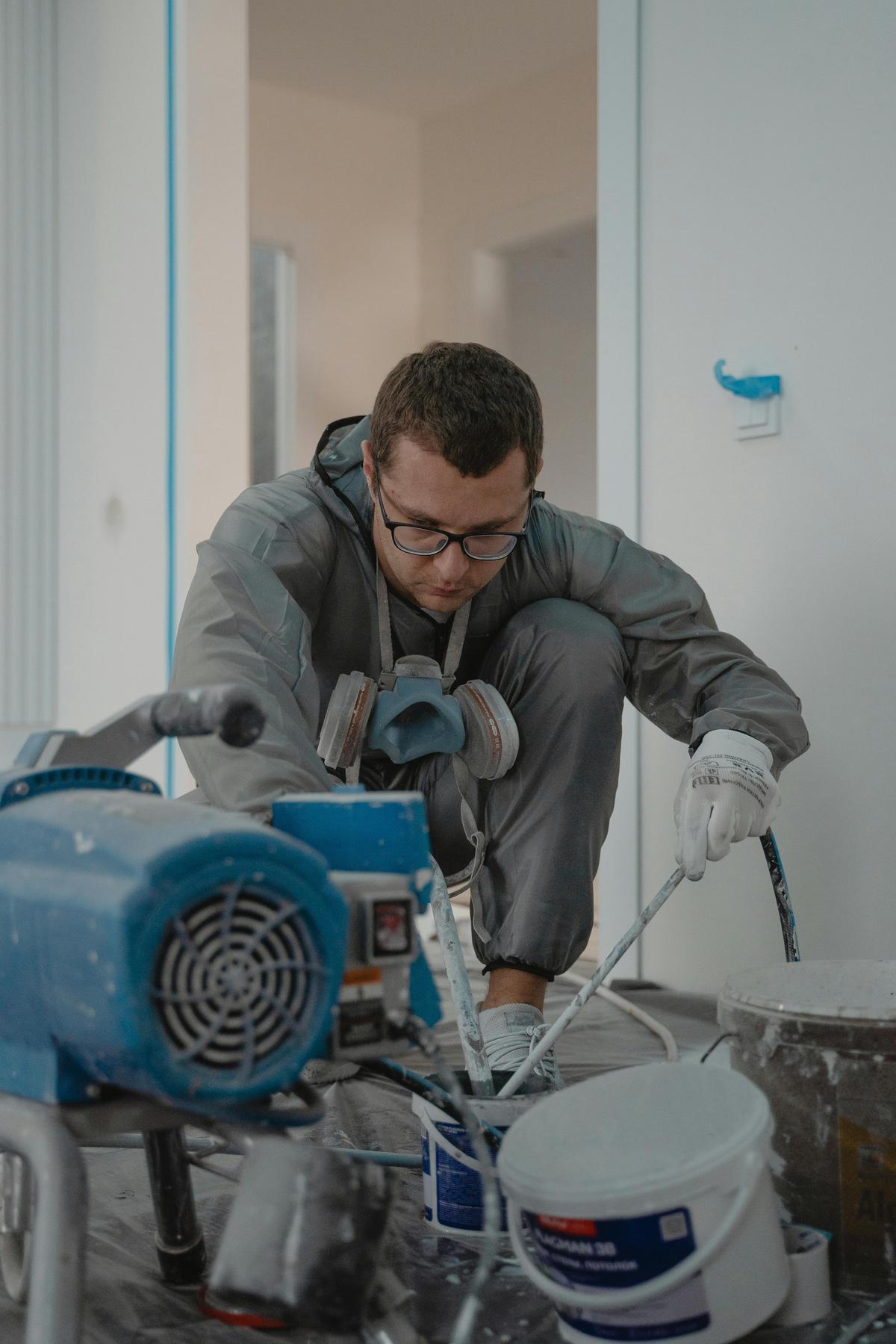 Worker in gray coveralls and a respirator crouched beside a paint sprayer pump in an unfinished room (sample photo)