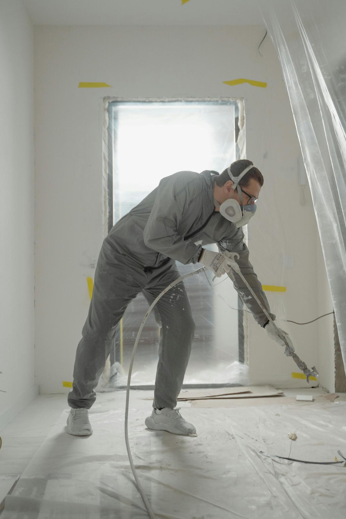 Worker in gray coveralls, a respirator and ear protection spraying inside a room covered with plastic sheeting (sample photo)