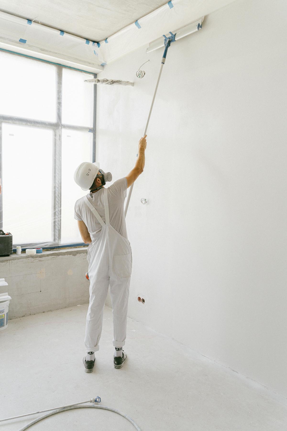 Worker in white coveralls and a head covering reaching up to roll paint on a wall and ceiling (sample photo)